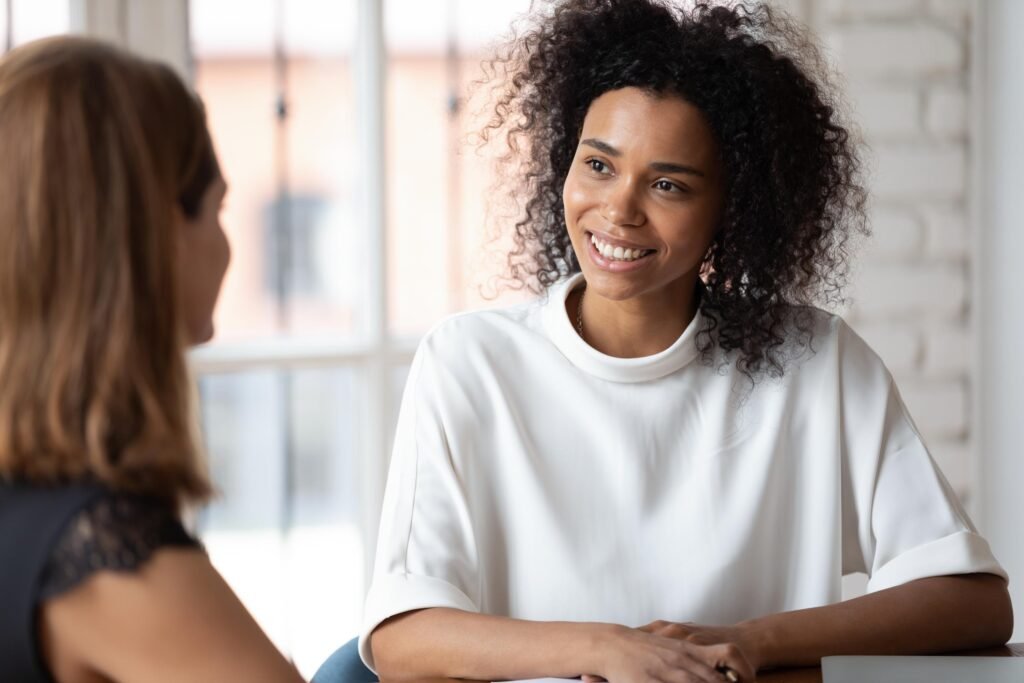 Two women engaged in a conversation during a holistic longevity consultation, one smiling and wearing a white top, in a bright, airy setting with natural light.