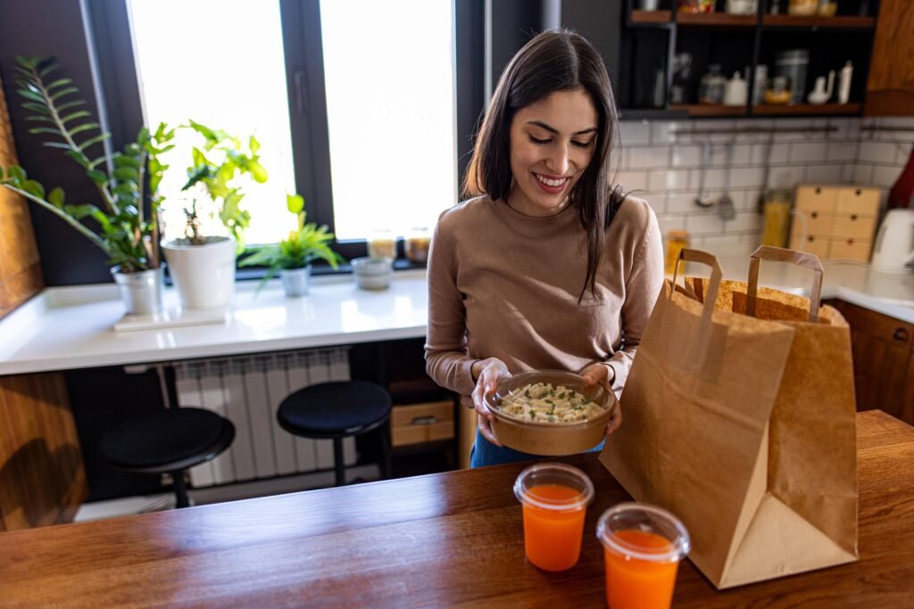 Woman smiling while holding a bowl of healthy food in a modern kitchen, with a paper bag and two orange drinks on the table, reflecting personalized wellness and convenience.